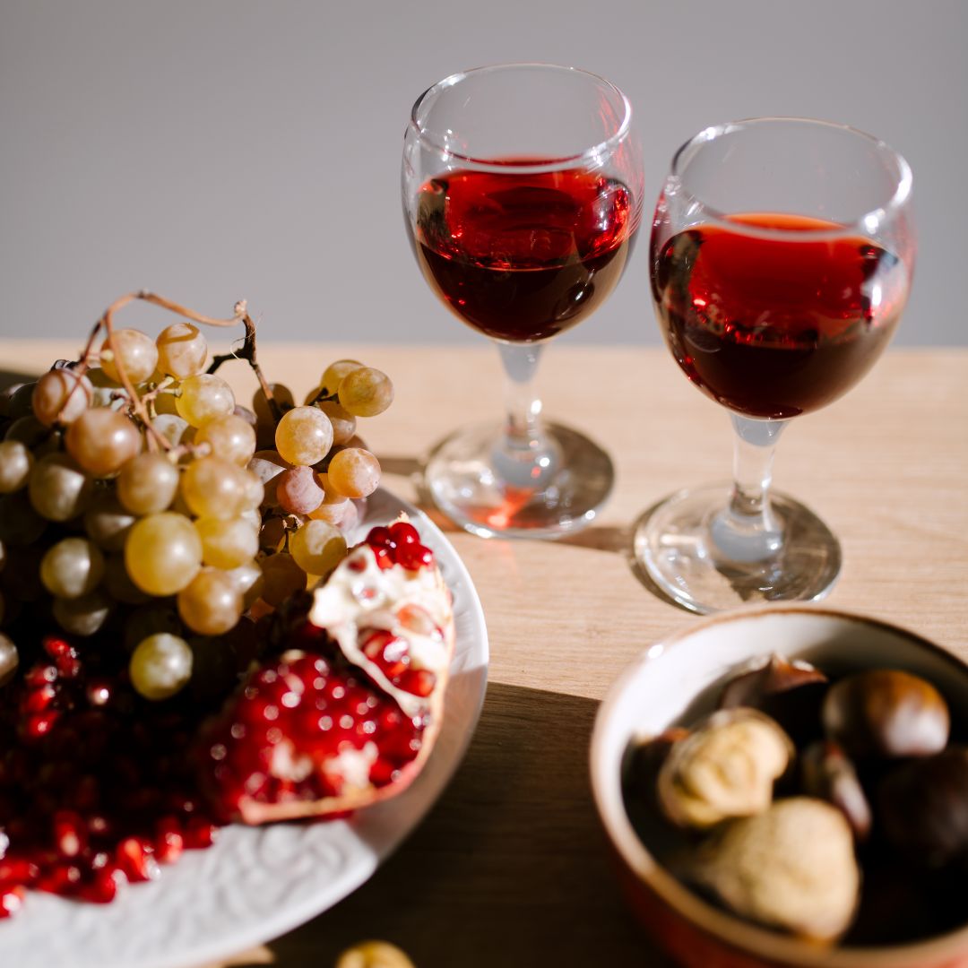 A glass of red wine on a wooden table next to a bowl of fresh red grapes, blueberries and a square of dark chocolate.